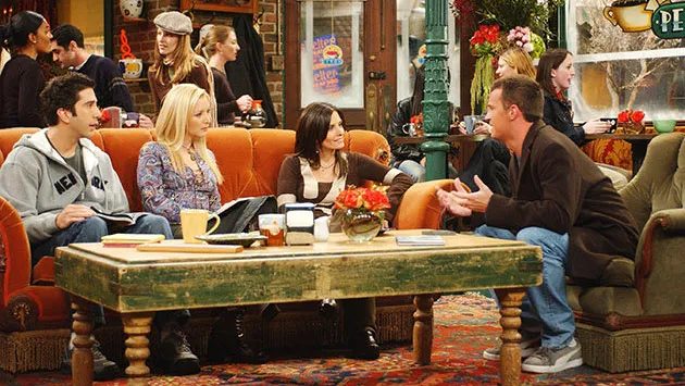 Sitcom still of six friends sitting together on sofas and armchairs around a low table in a coffee house.