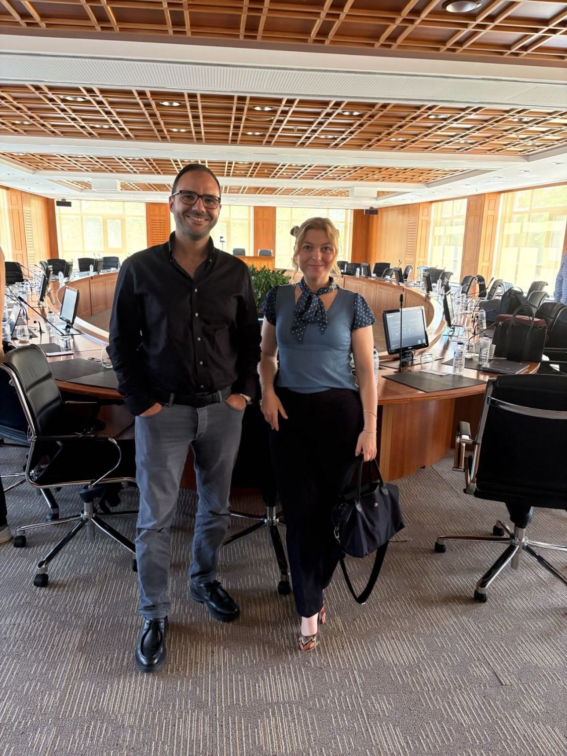 Two people standing and smiling in an empty wood-panelled boardroom with a large round conference table.
