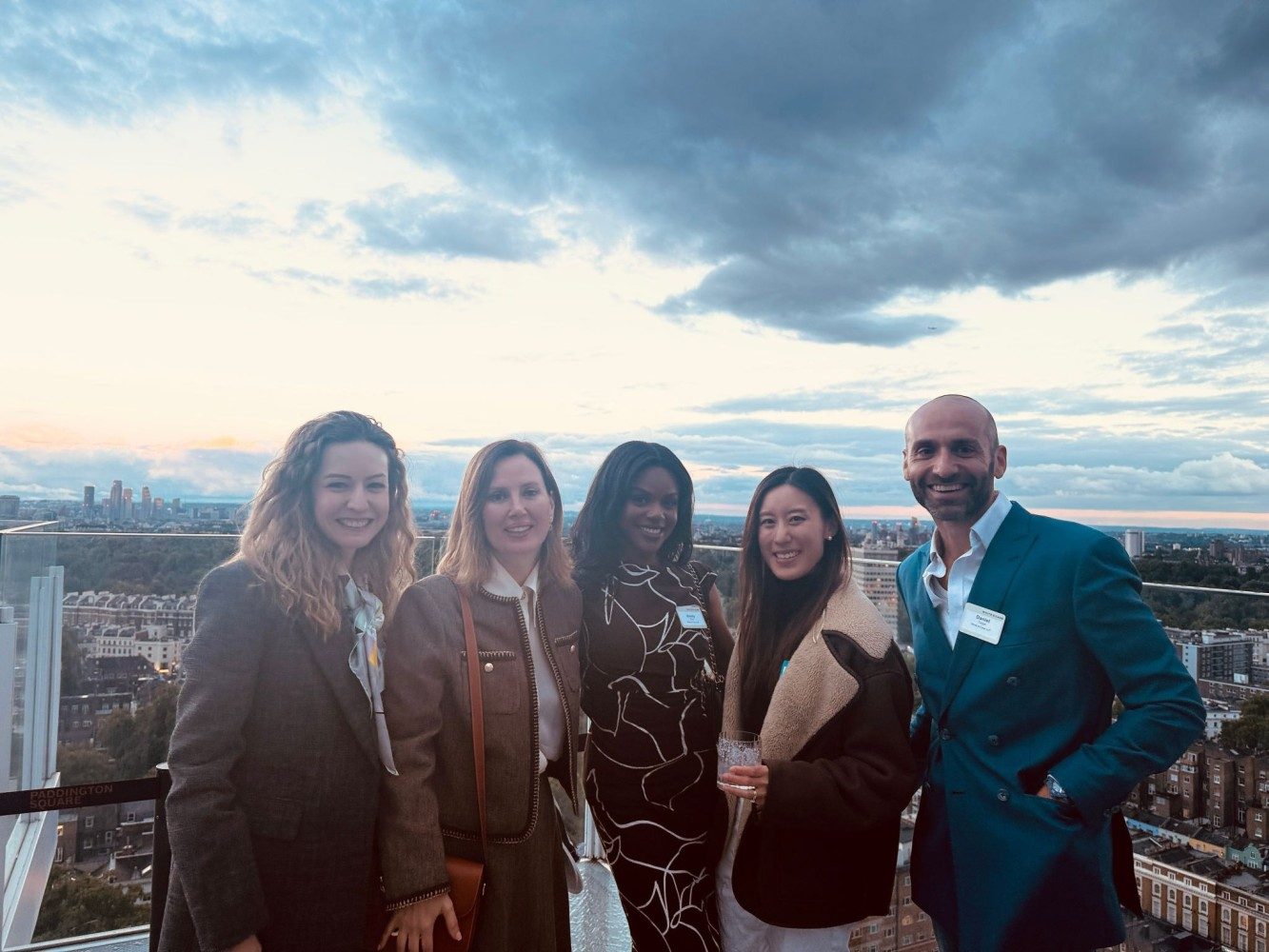 Five people at a rooftop reception in London at dusk, with the city skyline behind them.