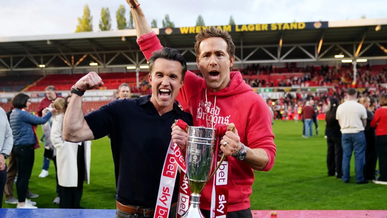 Two men celebrating on a football pitch, holding the Sky Bet League trophy in front of the Wrexham Lager Stand.
