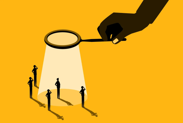 Illustration of a large hand holding a magnifying glass that casts a spotlight on one figure standing apart from a small crowd.