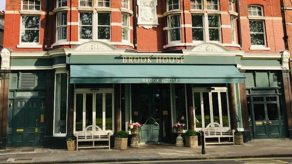 The green-painted frontage of Brook House, a red-brick London pub, with benches and flower baskets beside the door.