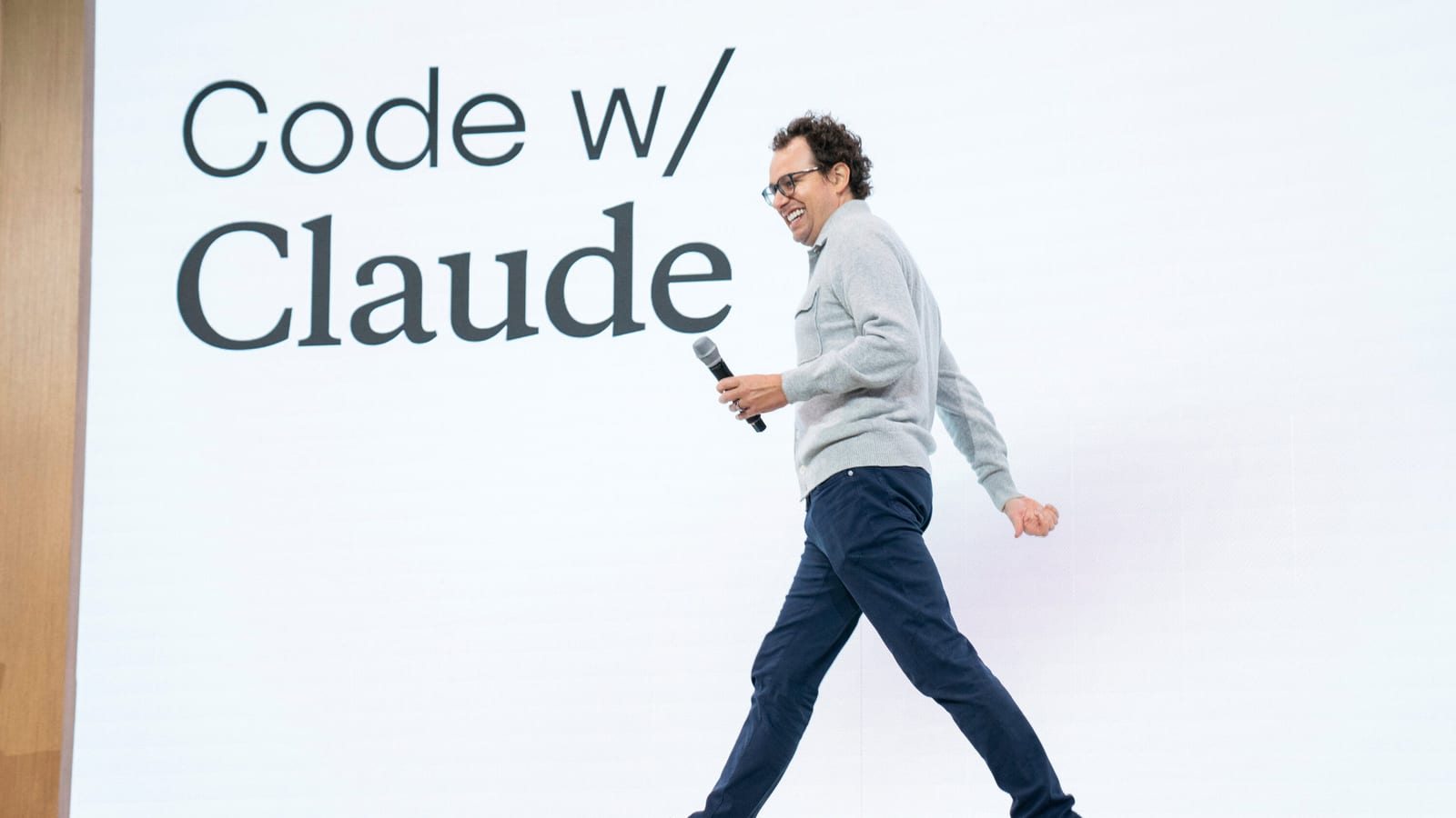 A speaker crossing a stage with a microphone in front of a large screen reading “Code w/ Claude”.